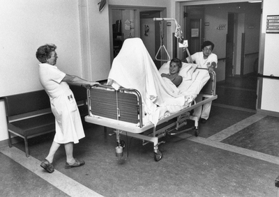 826716 Afbeelding van twee brancardiers die een patiënte in een ziekenhuisbed vervoeren in het Diakonessenhuis ...